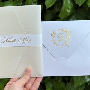 K&ouml;nnte beinhalten: Elfenbeinfarbene Hochzeitseinladung mit goldenem Folienmuster. Die Einladung zeigt die Namen "Daniella & Carlos" in goldener Schrift auf einem wei&szlig;en Band. Ein wei&szlig;er Umschlag mit goldenem Blumenschmuck ist ebenfalls zu sehen.