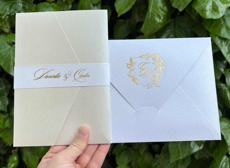 K&ouml;nnte beinhalten: Elfenbeinfarbene Hochzeitseinladung mit goldenem Folienmuster. Die Einladung zeigt die Namen "Daniella & Carlos" in goldener Schrift auf einem wei&szlig;en Band. Ein wei&szlig;er Umschlag mit goldenem Blumenschmuck ist ebenfalls zu sehen.