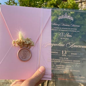 Pode incluir: Um convite de Quincea&ntilde;era em acr&iacute;lico rosa e transparente. O convite apresenta uma coroa, o texto "Marco y Veronica Amezcua" e o texto "Jacqueline Amezcua". O convite tamb&eacute;m inclui a data "Sabado 13 Julio 2024" e o local do evento.