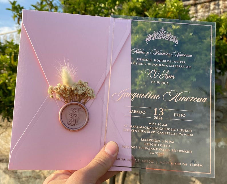 Pode incluir: Um convite de Quincea&ntilde;era em acr&iacute;lico rosa e transparente. O convite apresenta uma coroa, o texto "Marco y Veronica Amezcua" e o texto "Jacqueline Amezcua". O convite tamb&eacute;m inclui a data "Sabado 13 Julio 2024" e o local do evento.