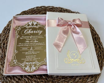 May include: A white box tied with a pink satin ribbon and bow, containing a gold-coloured invitation. The invitation has the word "Charity" at the top, with text below. The box is on a woven surface.