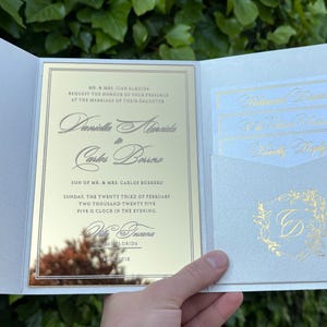 K&ouml;nnte beinhalten: Eine Hochzeitseinladungssuite mit einer verspiegelten goldenen Karte und einem hellblauen Au&szlig;enordner. Die Einladung enth&auml;lt die Namen des Paares, das Datum und die Uhrzeit der Zeremonie. Zus&auml;tzliche Karten befinden sich in einer Tasche.