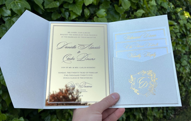 K&ouml;nnte beinhalten: Eine Hochzeitseinladungssuite mit einer verspiegelten goldenen Karte und einem hellblauen Au&szlig;enordner. Die Einladung enth&auml;lt die Namen des Paares, das Datum und die Uhrzeit der Zeremonie. Zus&auml;tzliche Karten befinden sich in einer Tasche.