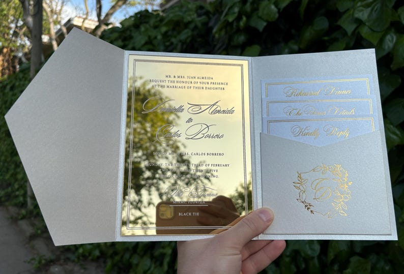 K&ouml;nnte beinhalten: Eine Hochzeitseinladung mit einer goldfarbenen Spiegelkarte und einem hellgrauen Au&szlig;enumschlag. Die Einladung enth&auml;lt die Namen des Paares und die Veranstaltungsdetails. Zus&auml;tzliche Karten sind in einer Tasche verstaut, und ein goldenes Blumendesign ist sichtbar.