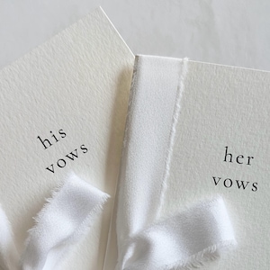 May include: Two off-white vow booklets with textured paper covers. The left booklet features the black printed text 'his vows' and the right booklet features 'her vows'. Each booklet is tied with a frayed white silk ribbon across the front cover.