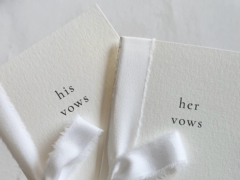 May include: Two off-white vow booklets with textured paper covers. The left booklet features the black printed text 'his vows' and the right booklet features 'her vows'. Each booklet is tied with a frayed white silk ribbon across the front cover.