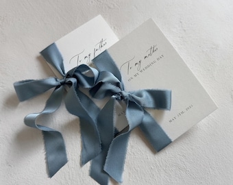 Puede incluir: Dos tarjetas rectangulares blancas con texto impreso en negro y cintas de seda deshilachadas de color azul polvoriento atadas en lazos. Una tarjeta dice To my father, la otra To my mother on my wedding day, May 5th, 2025. Las tarjetas se superponen.