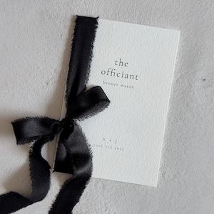 May include: A rectangular white card with a textured surface, featuring black printed text that reads the officiant, benner mason, s + j, and june 7th 2025. A black ribbon with frayed edges is tied vertically around the left side of the card.