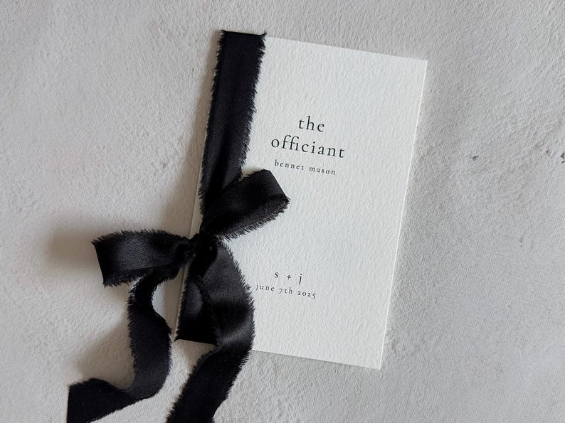 May include: A rectangular white card with a textured surface, featuring black printed text that reads the officiant, benner mason, s + j, and june 7th 2025. A black ribbon with frayed edges is tied vertically around the left side of the card.