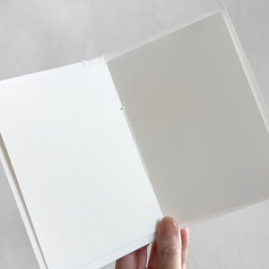 May include: A folded greeting card with a white exterior and a plain, light gray interior. The card is constructed from paper and features a simple, clean design with a visible center fold line, providing a blank space for writing messages.
