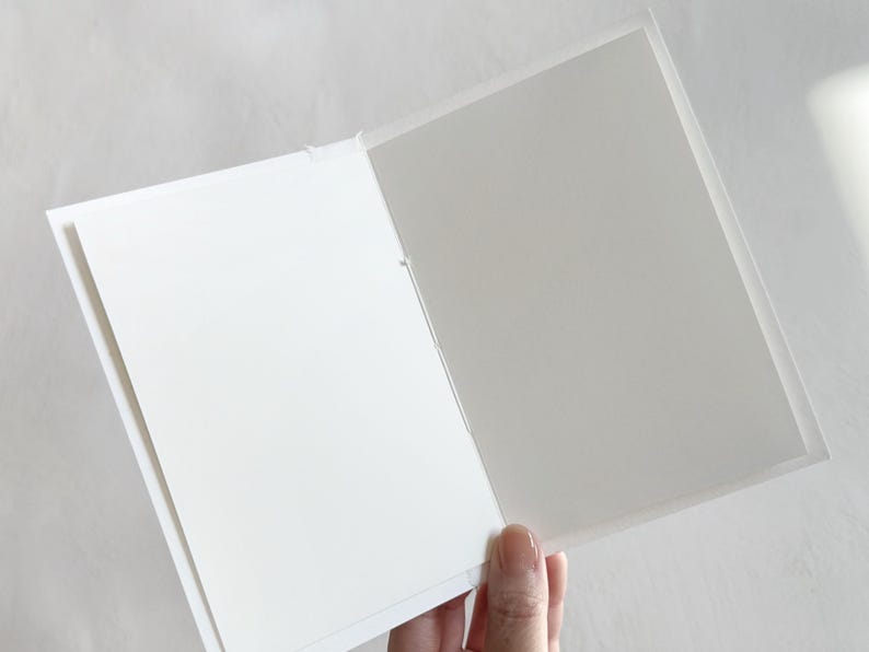 May include: A folded greeting card with a white exterior and a plain, light gray interior. The card is constructed from paper and features a simple, clean design with a visible center fold line, providing a blank space for writing messages.