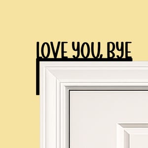 Black wood L-shaped door-corner signs reading “LOVE YOU, BYE” in bold letters. The Small version measures 10-3/4" wide × 6" tall, and the Large version measures 17-1/4" wide × 7-3/8" tall. Both are 1/4" thick and designed to perch on a door-frame corner without installation hardware.