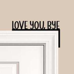 Black wood L-shaped door-corner signs reading “LOVE YOU, BYE” in bold letters. The Small version measures 10-3/4" wide × 6" tall, and the Large version measures 17-1/4" wide × 7-3/8" tall. Both are 1/4" thick and designed to perch on a door-frame corner without installation hardware.
