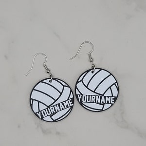 May include: A pair of silver earrings with white volleyball-shaped charms. The charms have black outlines and the text "YOURNAME" printed on them.