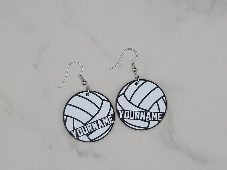 May include: A pair of silver earrings with white volleyball-shaped charms. The charms have black outlines and the text "YOURNAME" printed on them.