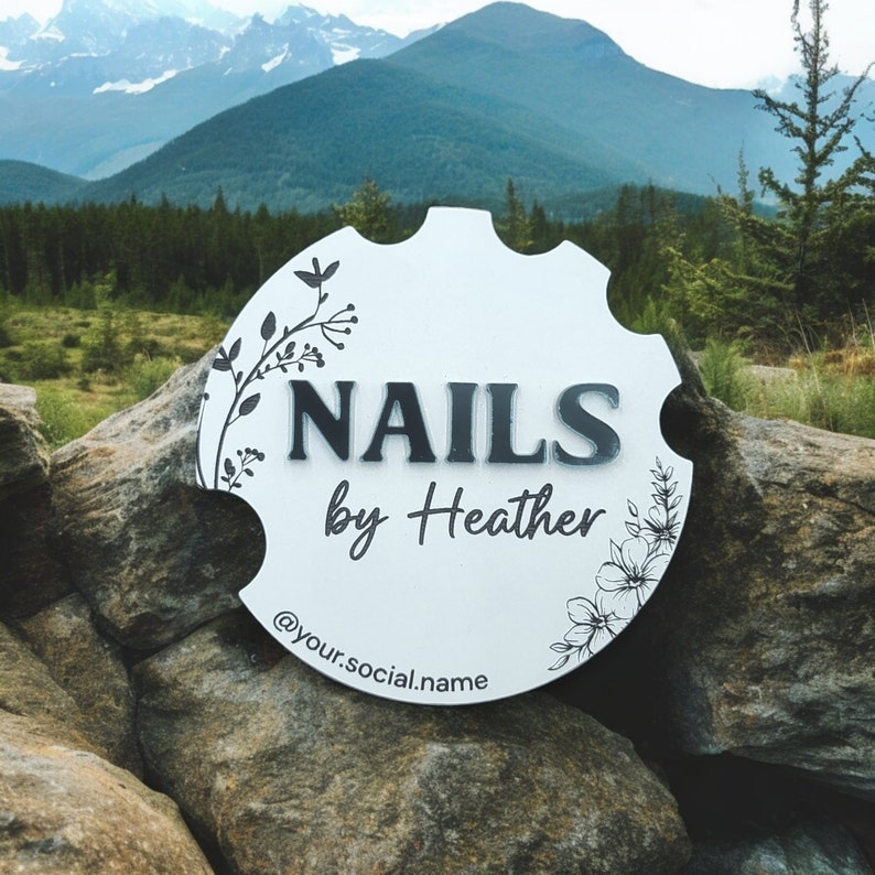 May include: White round sign with black floral designs and the text "NAILS by Heather" and the social media handle "@your.social.name".