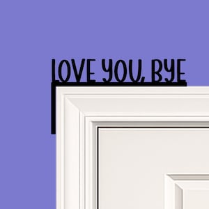 Black wood L-shaped door-corner signs reading “LOVE YOU, BYE” in bold letters. The Small version measures 10-3/4" wide × 6" tall, and the Large version measures 17-1/4" wide × 7-3/8" tall. Both are 1/4" thick and designed to perch on a door-frame corner without installation hardware.