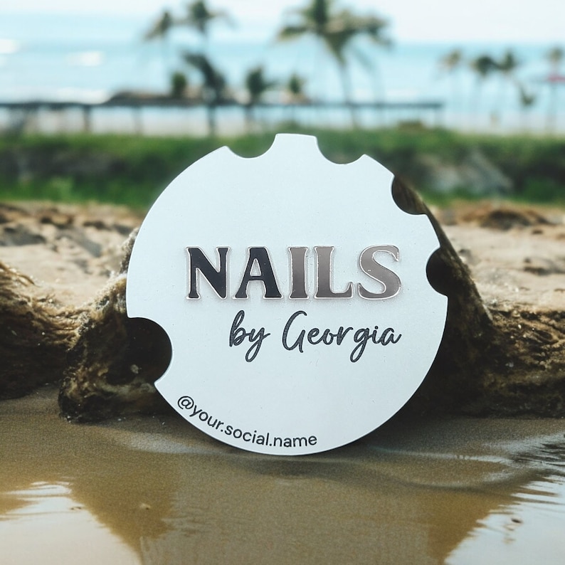 May include: White round sign with the text "NAILS by Georgia" and the social media handle "@your.social.name" on a sandy beach.