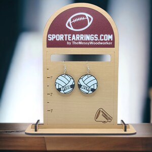 May include: A pair of personalized volleyball earrings with the name "Averie" on each earring. The earrings are displayed on a wooden stand with a ruler for size reference. The stand also features the text "SPORTEARRINGS.COM by TheMessyWoodworker" and a small image of a megaphone with the text "PERSONALIZED EARRINGS".