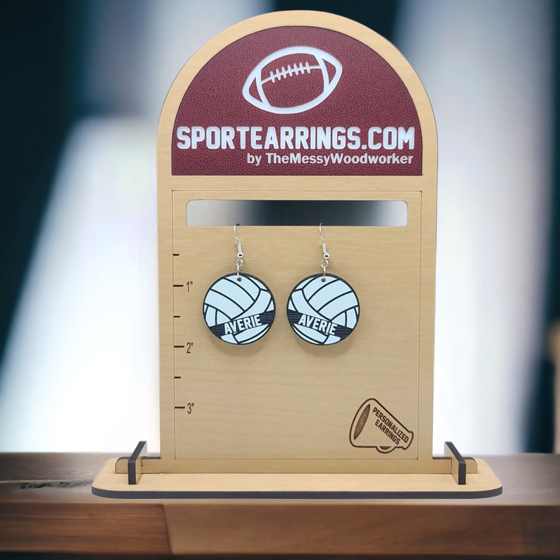 May include: A pair of personalized volleyball earrings with the name "Averie" on each earring. The earrings are displayed on a wooden stand with a ruler for size reference. The stand also features the text "SPORTEARRINGS.COM by TheMessyWoodworker" and a small image of a megaphone with the text "PERSONALIZED EARRINGS".