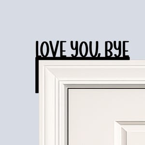 Black wood L-shaped door-corner signs reading “LOVE YOU, BYE” in bold letters. The Small version measures 10-3/4" wide × 6" tall, and the Large version measures 17-1/4" wide × 7-3/8" tall. Both are 1/4" thick and designed to perch on a door-frame corner without installation hardware.
