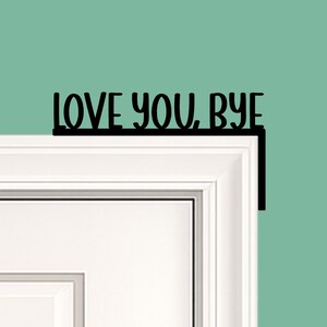 Black wood L-shaped door-corner signs reading “LOVE YOU, BYE” in bold letters. The Small version measures 10-3/4" wide × 6" tall, and the Large version measures 17-1/4" wide × 7-3/8" tall. Both are 1/4" thick and designed to perch on a door-frame corner without installation hardware.