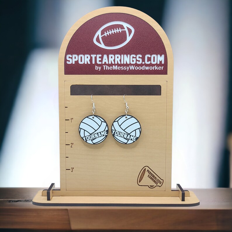May include: A pair of personalized volleyball earrings on a wooden display stand. The earrings are white with black stitching and the text "YOURNAME" is printed on each earring. The stand has a ruler on the side and the text "SPORTEARRINGS.COM by TheMessyWoodworker" at the top.