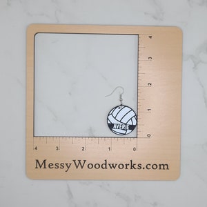 May include: A pair of white and black volleyball earrings with the name "Averie" printed on the front. The earrings are shown on a wooden ruler with a white background.