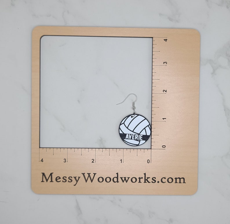 May include: A pair of white and black volleyball earrings with the name "Averie" printed on the front. The earrings are shown on a wooden ruler with a white background.
