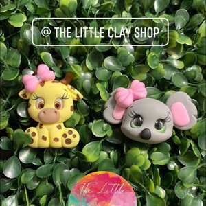 Puede incluir: Dos figuras de animales de arcilla hechas a mano: una jirafa amarilla con manchas marrones y un koala gris, ambos con lazos rosas. El fondo es un lecho de hojas verdes. El texto "@ THE LITTLE CLAY SHOP" está en la parte superior y un logotipo colorido con el mismo texto en la parte inferior.