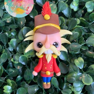 Puede incluir: Una figurita de cascanueces hecha a mano de arcilla polimérica con un uniforme rojo y dorado, una barba amarilla y una pluma roja en su sombrero. La figurita está sentada sobre un lecho de hojas verdes.