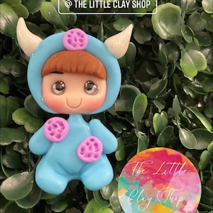 Puede incluir: Una figurita de arcilla azul de un niño que lleva un disfraz de monstruo azul con manchas moradas. La figurita tiene el pelo castaño, una cara sonriente y ojos grandes. La figurita lleva un disfraz de monstruo azul con un cuerno blanco a cada lado de la cabeza. La figurita está de pie sobre un fondo verde frondoso. El texto "The Little Clay Shop" es visible en la imagen.