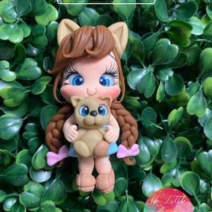 Puede incluir: Una figurita hecha a mano de arcilla polimérica de una niña con cabello castaño, vestida con un vestido azul y sosteniendo un cachorro marrón. La niña tiene ojos azules y lleva lazos rosas en el cabello. La figurita está colocada sobre un fondo verde de hojas. El texto "@ THE LITTLE CLAY SHOP" es visible en la parte superior de la imagen.