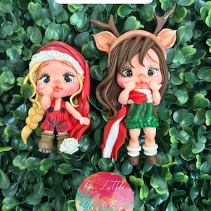 Puede incluir: Dos figuras de arcilla hechas a mano. Una está vestida con un traje de Papá Noel rojo con barba y gorro blancos. La otra es un reno con pelo castaño, cuernos y una bufanda roja y blanca. The Little Clay Shop.