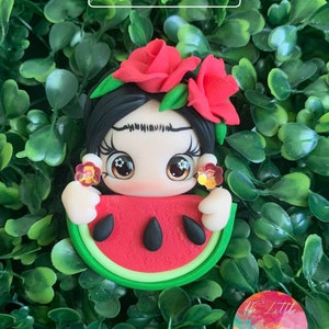May include: A handmade polymer clay figurine of a woman with black hair, wearing a red flower crown and holding a slice of watermelon. The figurine has large eyes and a whimsical expression. The background is a green leafy surface.