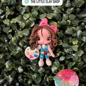 Puede incluir: Figura de arcilla hecha a mano de una joven artista con cabello castaño rizado y un lazo rosa. Lleva un peto azul y sostiene un pincel y una paleta con pintura colorida. El logotipo de The Little Clay Shop es visible.