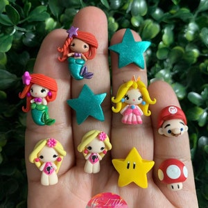 Puede incluir: Una colección de encantos de arcilla coloridos con personajes de videojuegos y películas populares. Los encantos incluyen una sirena, una princesa, una estrella, un hongo y un personaje del videojuego Super Mario Bros. Los encantos están hechos a mano y serían perfectos para agregar un toque de fantasía a cualquier proyecto.
