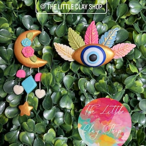 May include: Two handmade clay charms, one a crescent moon with floral details and dangling charms, the other a blue evil eye with colorful feathers. The charms are on a green leafy background with a sticker that says "The Little Clay Shop".