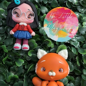 Puede incluir: Una figurita de arcilla hecha a mano de una niña con cabello negro, que lleva gafas, una chaqueta roja, una falda azul y zapatos rosas. Está de pie junto a una figurita de arcilla hecha a mano de un panda rojo con orejas blancas y vientre blanco. El fondo es una superficie verde frondosa. El texto "@ THE LITTLE CLAY SHOP" es visible en la esquina superior derecha de la imagen.