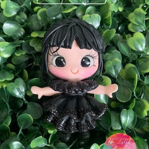 Puede incluir: Una muñeca negra y rosa con cabello negro y un vestido negro brillante. La muñeca lleva zapatos negros y tiene una gran sonrisa. La muñeca está sentada sobre un fondo verde.