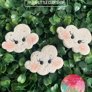 Puede incluir: Tres nubes de arcilla blanca con purpurina y detalles pintados. Cada nube tiene una cara sonriente con ojos negros y pestañas. Las nubes están dispuestas sobre un fondo de hojas verdes. El texto "@ THE LITTLE CLAY SHOP" es visible en la parte superior de la imagen.