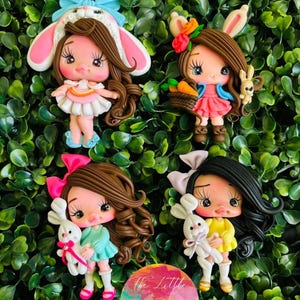 Puede incluir: Cuatro figuritas de arcilla polimérica hechas a mano de niñas vestidas con atuendos de Pascua. Cada niña tiene el pelo castaño, ojos grandes y sostiene un conejito. Las niñas llevan vestidos rosas, azules, amarillos y blancos. El fondo es un arbusto verde frondoso.