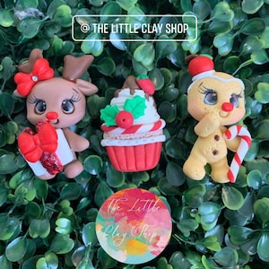 Puede incluir: Tres adornos de arcilla con temática navideña: un reno con un lazo rojo y un regalo, un cupcake con glaseado rojo y blanco y un hombre de jengibre con un bastón de caramelo.
