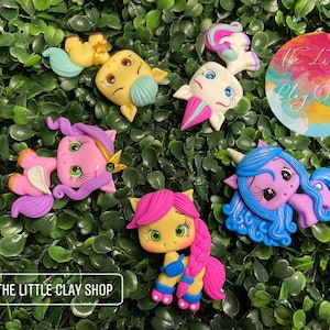 Puede incluir: Cinco figuritas de unicornio de colores con diferentes colores y estilos de pelo. Los unicornios están hechos de arcilla polimérica y tienen detalles pintados. Los unicornios están sobre un fondo verde.