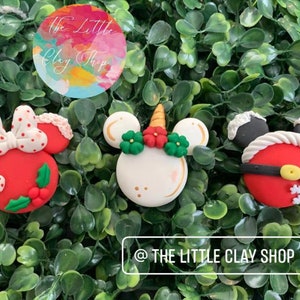 Puede incluir: Tres adornos navideños de arcilla con una Minnie Mouse de lunares rojos y blancos, un unicornio blanco con un cuerno dorado y acebo rojo y verde, y un Mickey Mouse Papá Noel rojo y blanco.