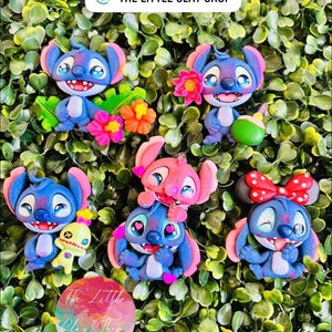 May include: A collection of handmade clay figurines featuring the character Stitch in various poses. The figurines are primarily blue and pink, with details like flowers, bows, and small accessories. The background is a bed of green leaves.
