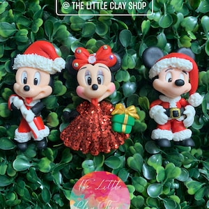 Puede incluir: Tres figuras de arcilla con temática navideña de Mickey Mouse, Minnie Mouse y Goofy. Mickey y Goofy llevan gorros de Papá Noel y atuendos rojos y blancos. Minnie lleva un vestido rojo brillante y un lazo rojo con pedrería. Las figuras están sobre un fondo verde con el texto "@ THE LITTLE CLAY SHOP".