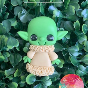 Puede incluir: Una figurita hecha a mano de arcilla polimérica de un personaje popular de la franquicia Star Wars. El personaje es verde y lleva un abrigo de piel y tiene ojos grandes.