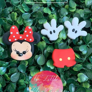 May include: A set of four clay charms featuring a red and white Minnie Mouse head, a pair of white Mickey Mouse gloves, and a pair of red Mickey Mouse shorts with yellow buttons. The charms are on a green background with the text "@ THE LITTLE CLAY SHOP" and "The Little Clay Shop".