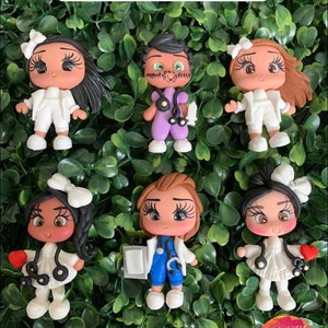 Puede incluir: Conjunto de seis figuritas de médicos en miniatura hechas de arcilla polimérica. Las figuritas están vestidas con batas blancas y uniformes médicos con estetoscopios. Las figuritas tienen diferentes peinados y rasgos faciales. Las figuritas están dispuestas sobre un fondo verde frondoso. El texto "@THE LITTLE CLAY SHOP" es visible en la parte superior de la imagen.